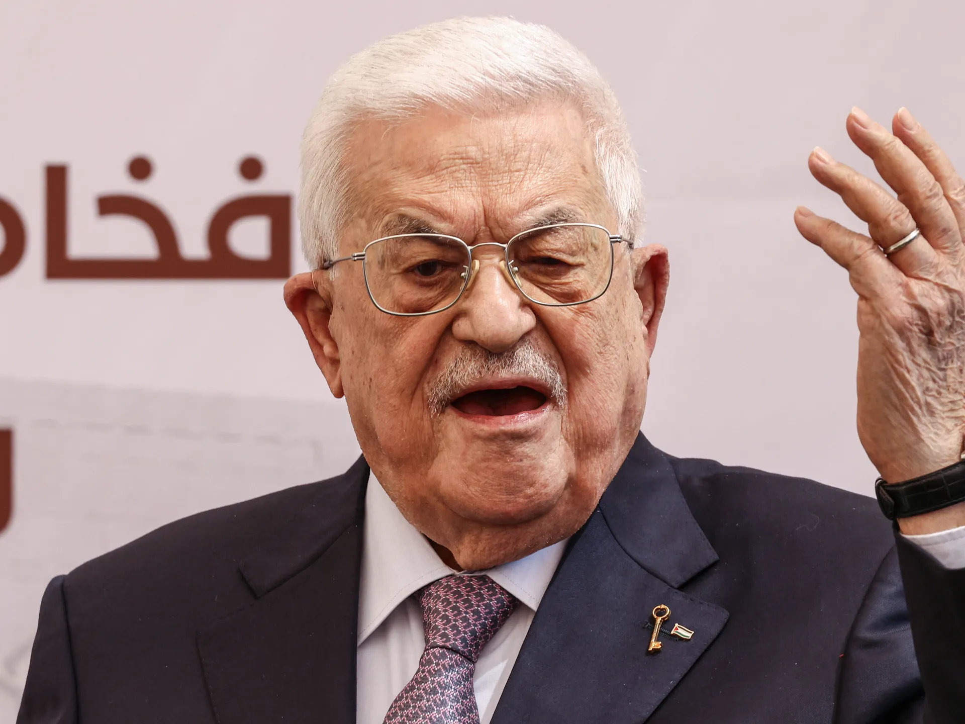 Elderly man wearing glasses and a suit speaks with one hand raised.