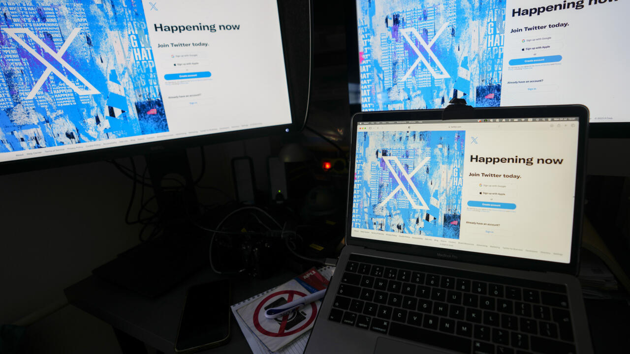 A laptop and larger monitor display X's blue login page in a dim workspace.