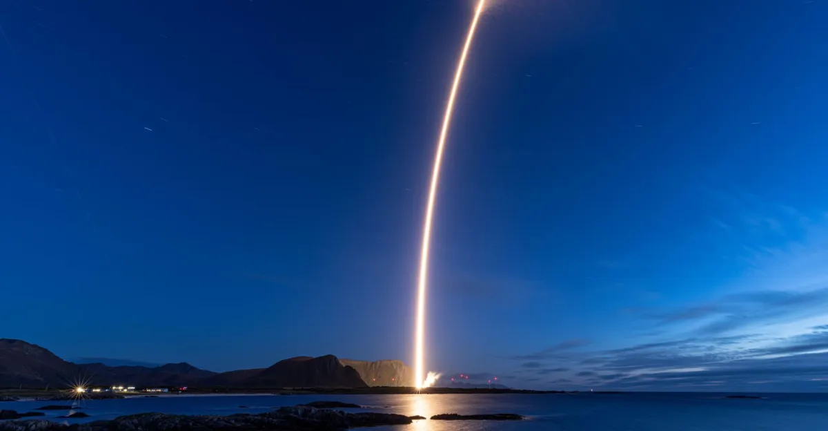 Rocket launches vertically over a dark coastal landscape, casting a bright reflection across calm water.