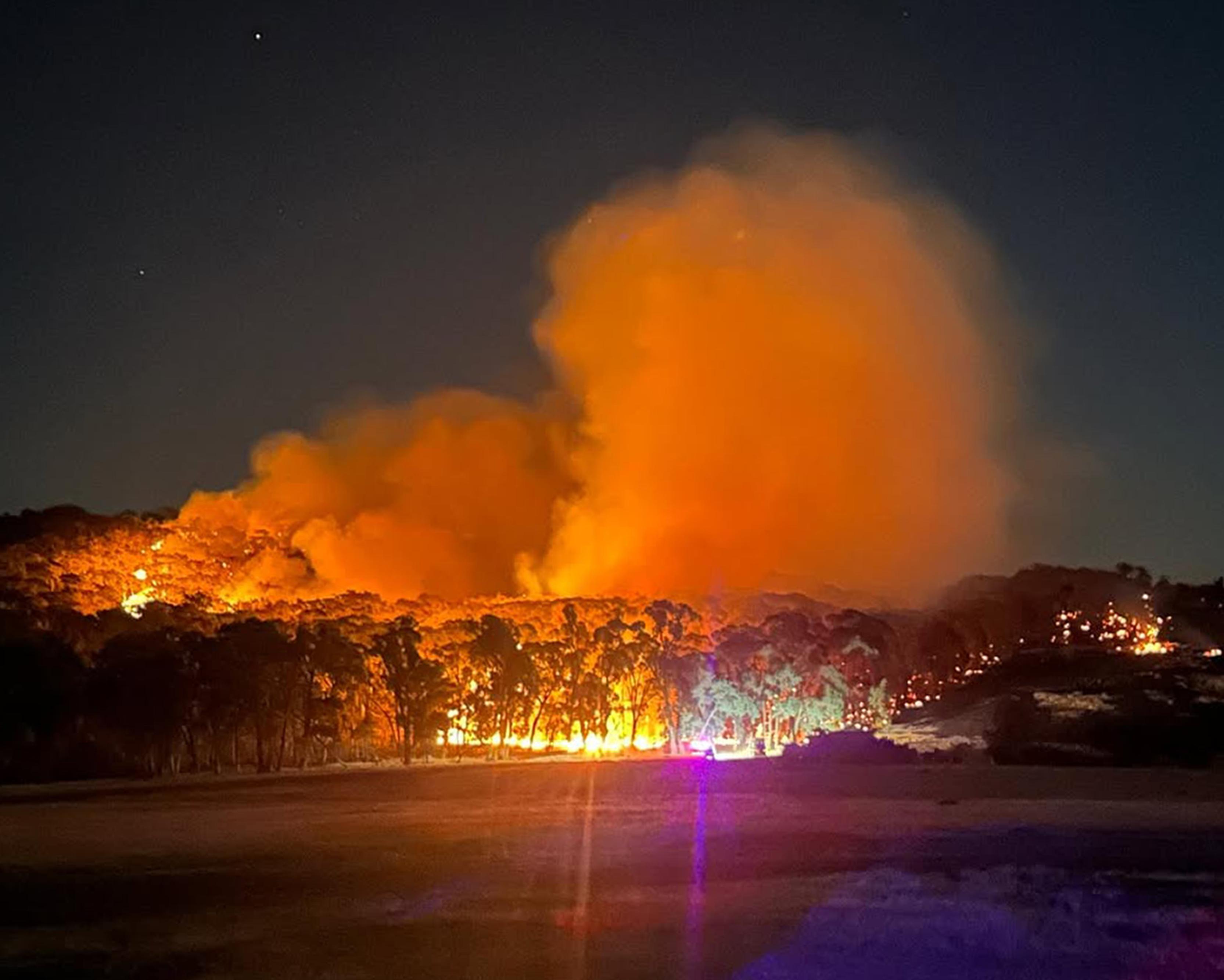 Nighttime wildfire burns through vegetation, sending a huge orange plume of smoke into the sky.