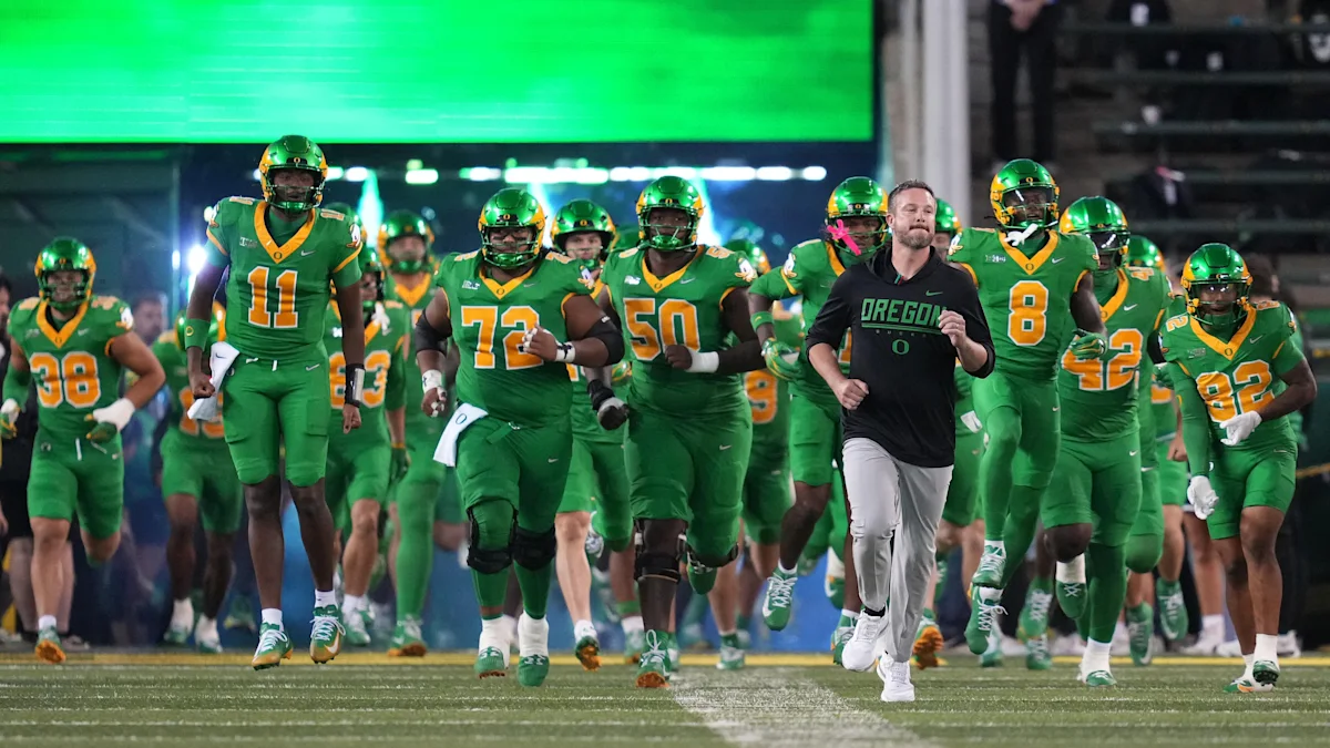 Oregon football players and a coach run onto the field beneath a bright green stadium display.