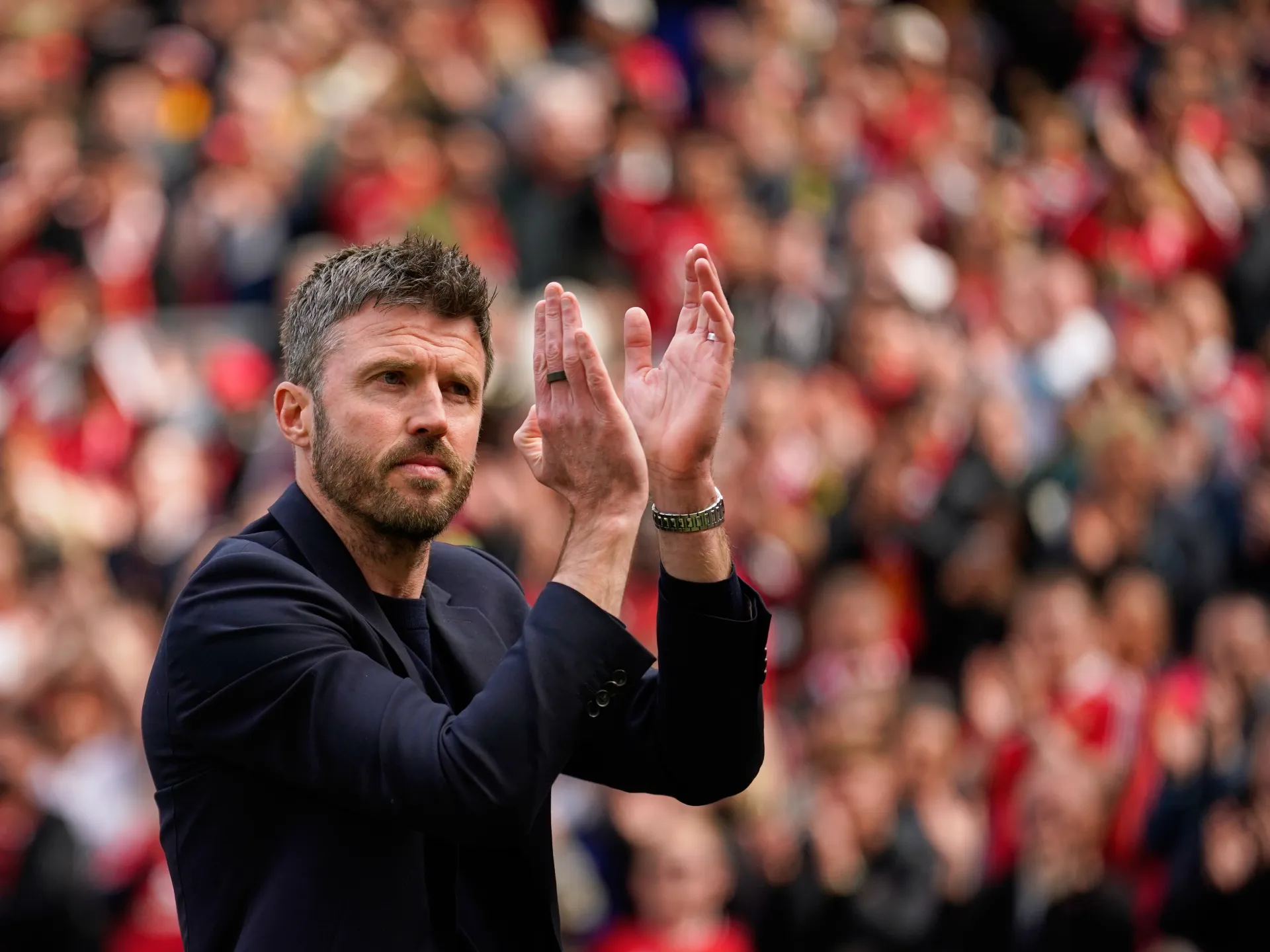 Bearded man in dark jacket claps before a blurred crowd at a football stadium