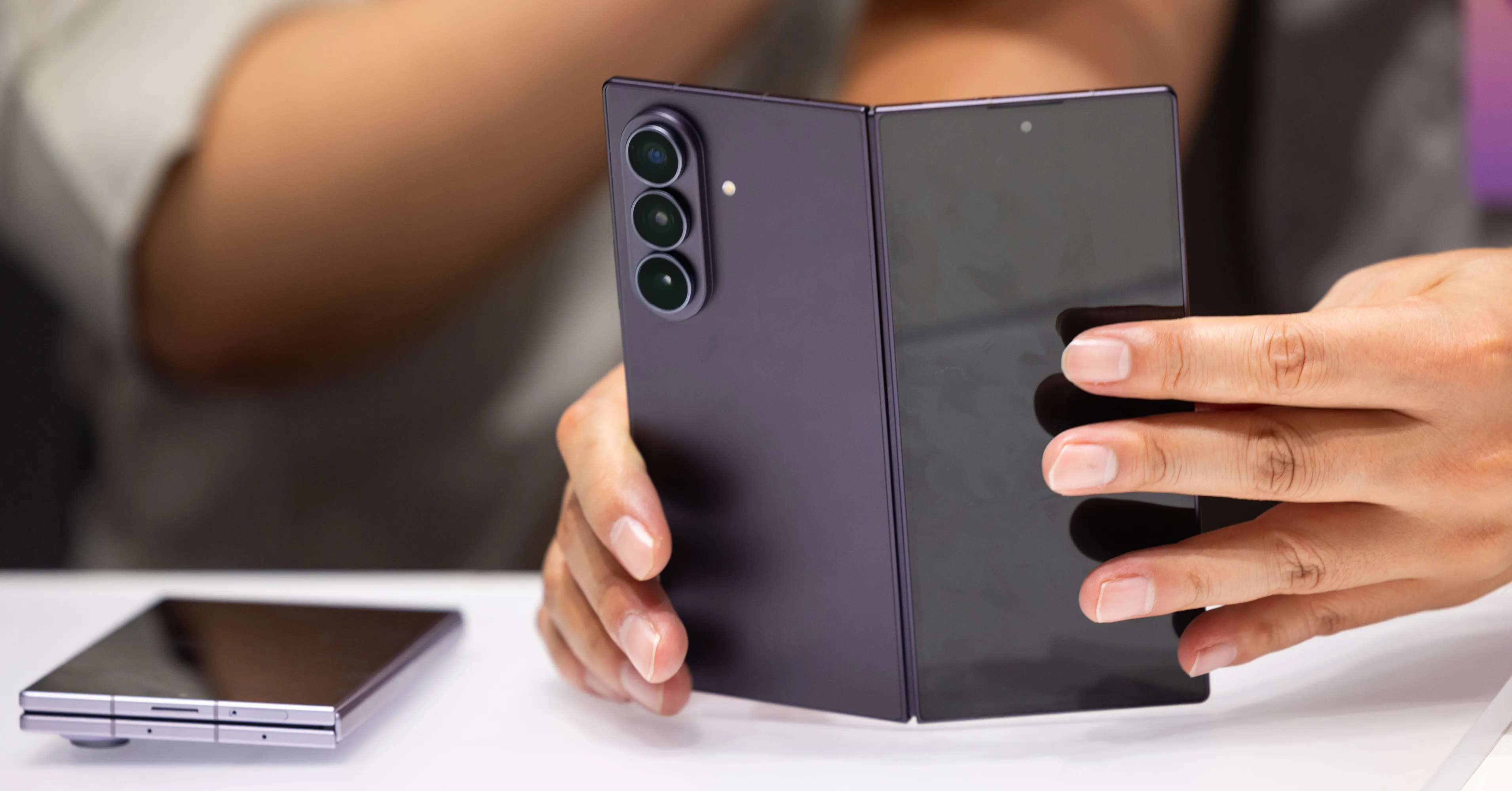 Person holds an open foldable smartphone beside another folded device on a white table.