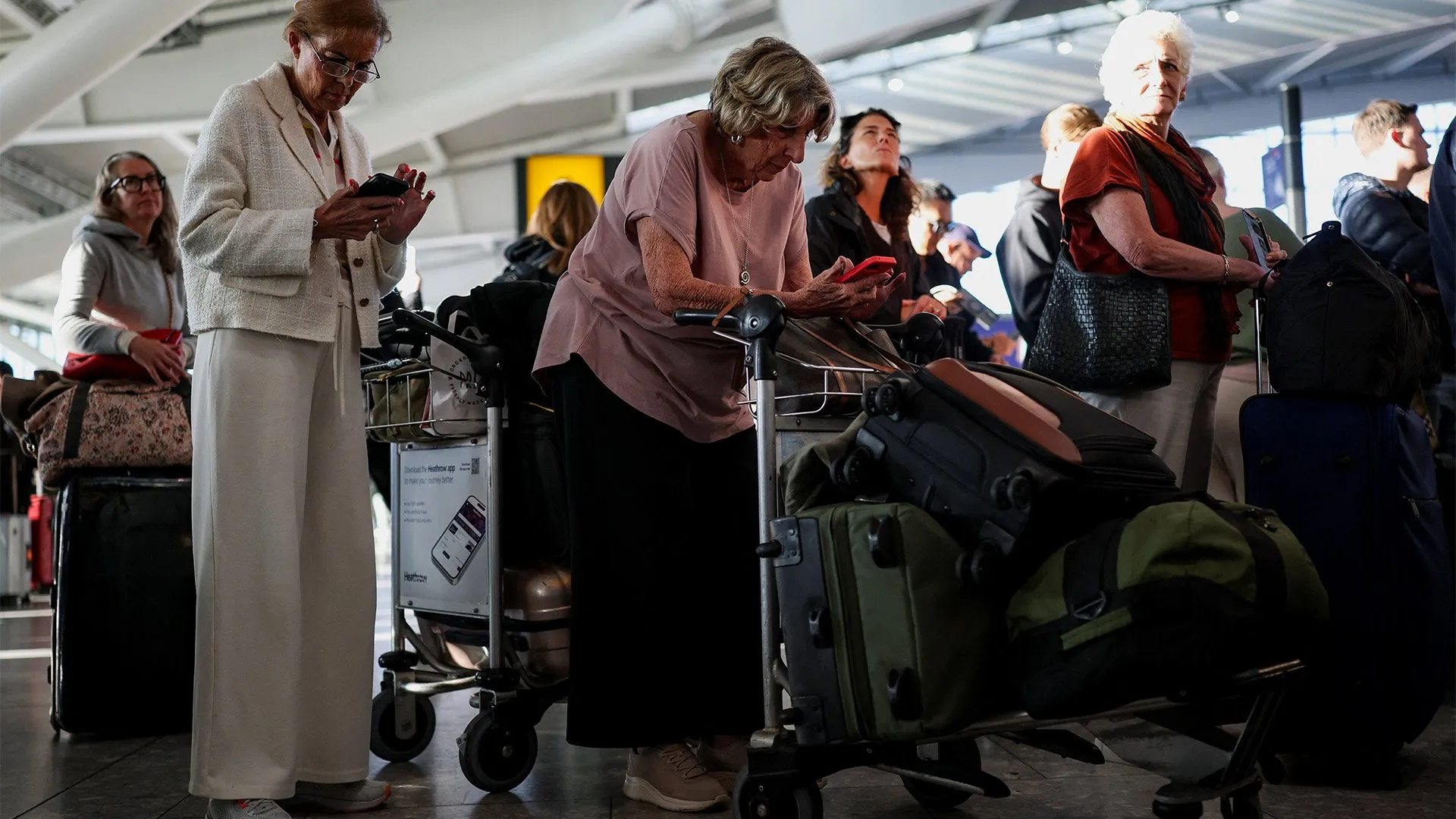 Travellers with luggage trolleys use smartphones in a crowded airport terminal.