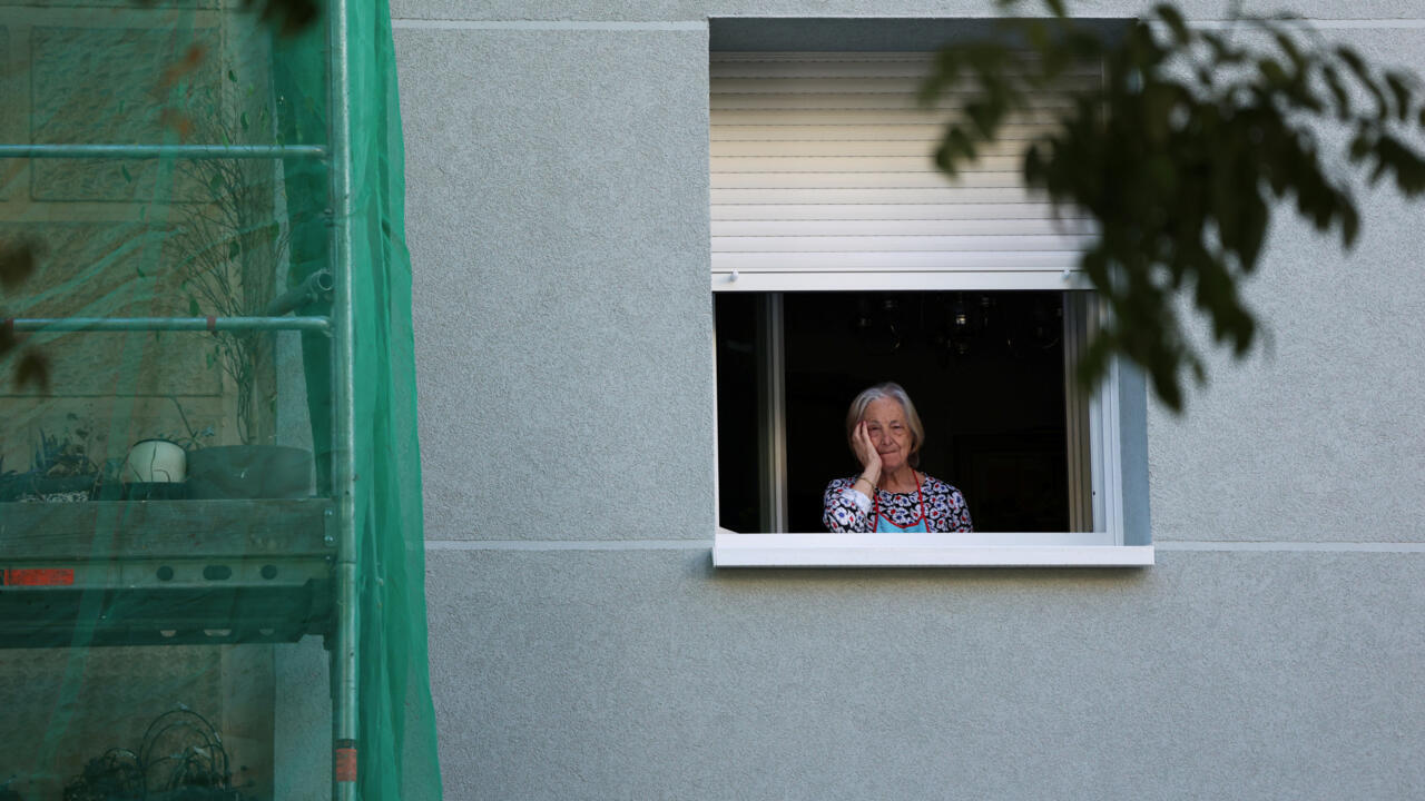 An elderly woman stands at an open window in a gray building, partly obscured by foreground leaves.