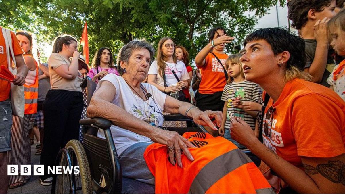An elderly woman in a wheelchair holds an orange vest as a volunteer crouches beside her at a gathering.
