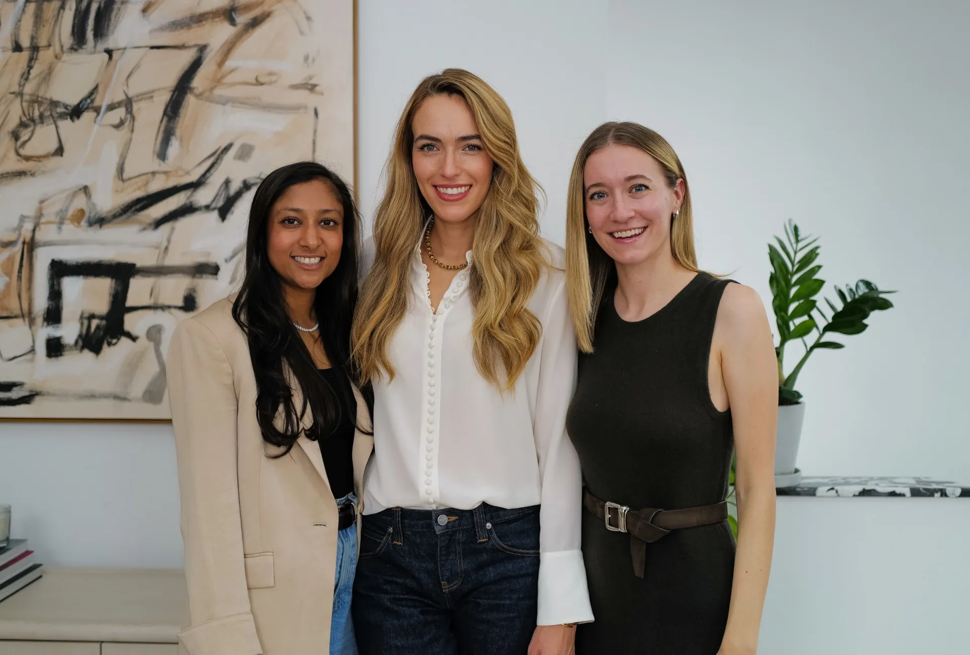 Three women pose together indoors beside abstract artwork and a potted plant.