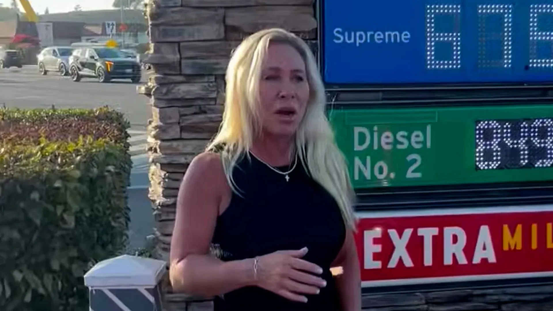 Blonde woman gestures beside a gas station price sign displaying Supreme and Diesel prices.