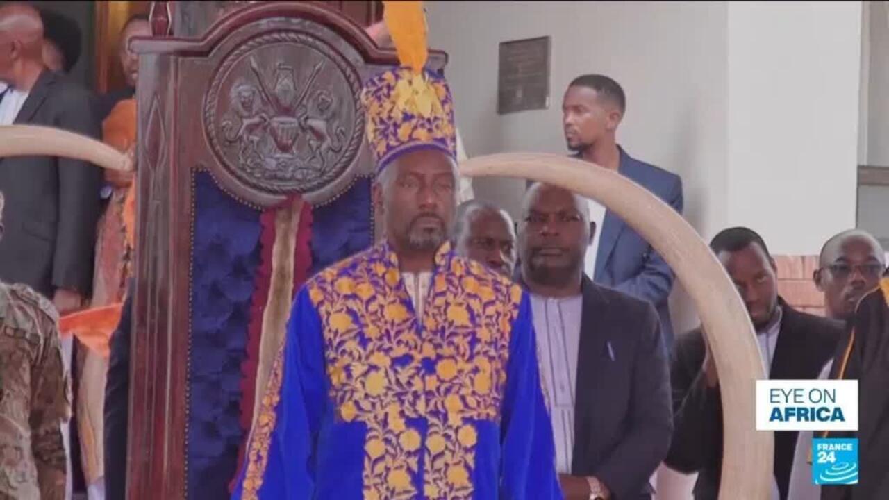 A man in blue and gold ceremonial clothing stands before an ornate throne, surrounded by attendees.