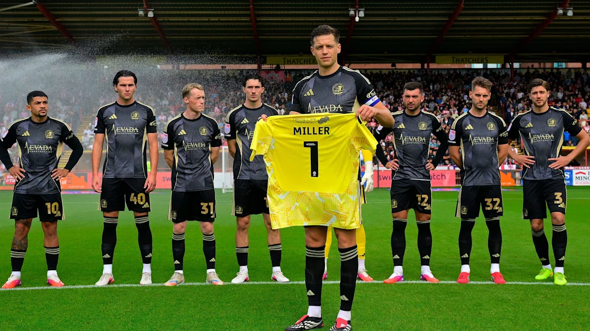 Football team members stand on pitch while goalkeeper holds a yellow shirt bearing Miller and number one