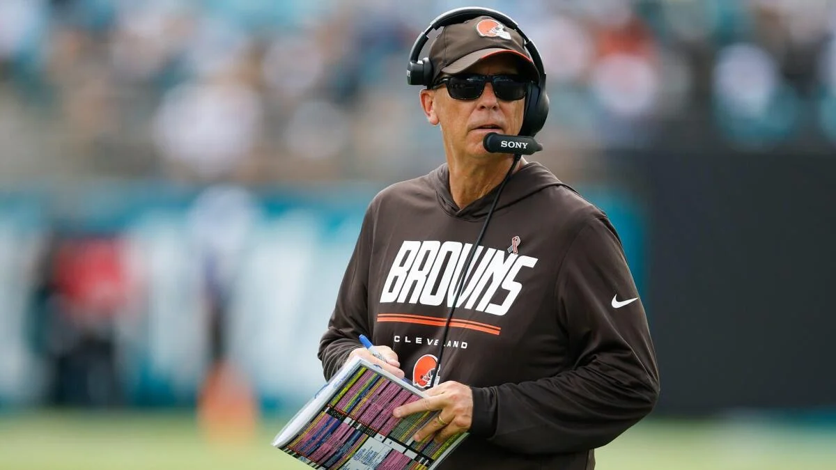 Cleveland Browns coach wearing headset and sunglasses holds a play sheet on the sideline