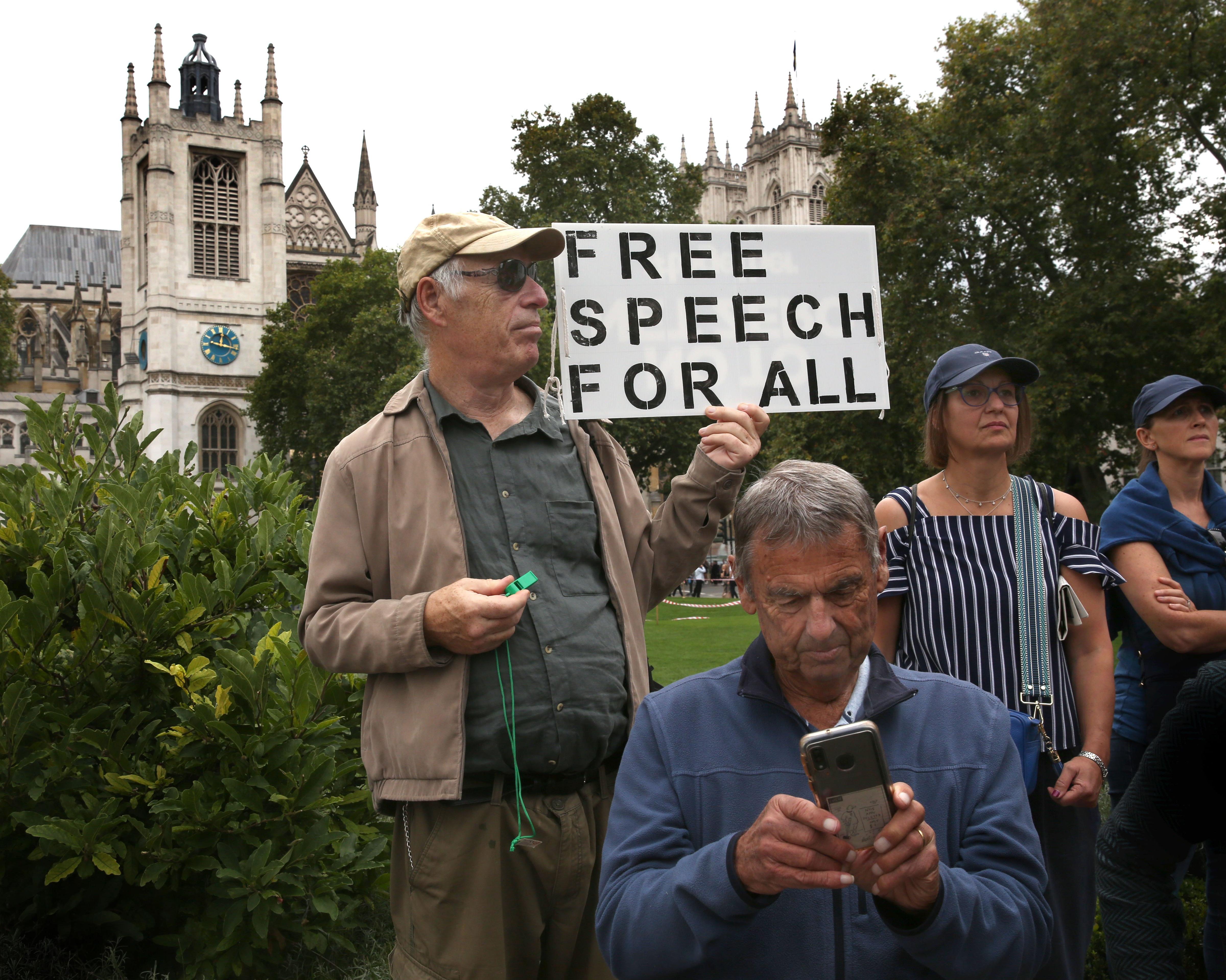 A man holds a “Free Speech for All” sign among people outside a Gothic church.