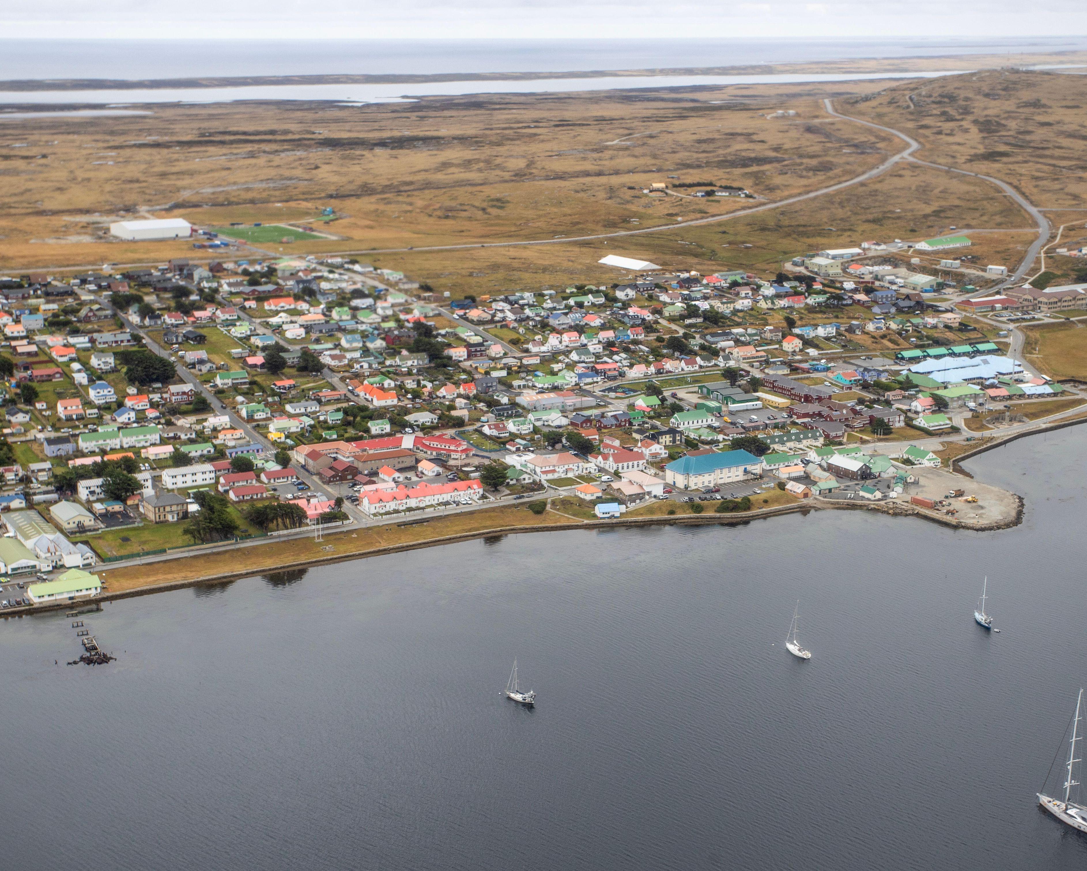 Aerial view of a colorful coastal settlement beside calm water, roads, fields and several sailboats.