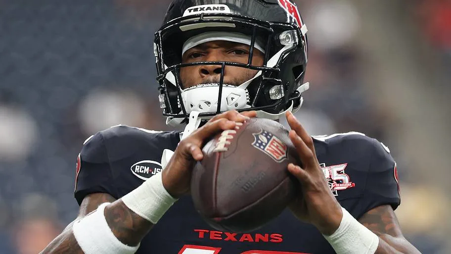 Houston Texans player in a dark uniform and helmet holds a football in a passing stance.