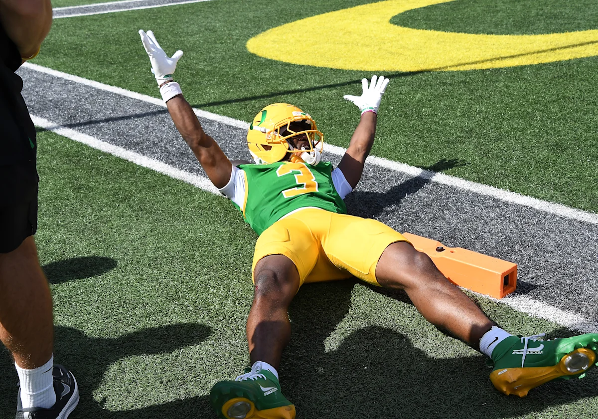 College football player in green and yellow uniform lies on turf with arms raised near end zone.