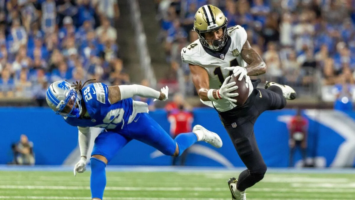 Football player in white and black uniform hurdles a blue-clad defender while securing the ball.