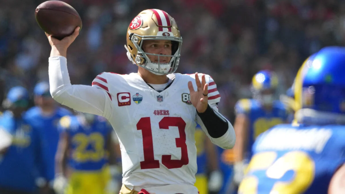 Quarterback in a San Francisco 49ers uniform raises a football to throw during a game.