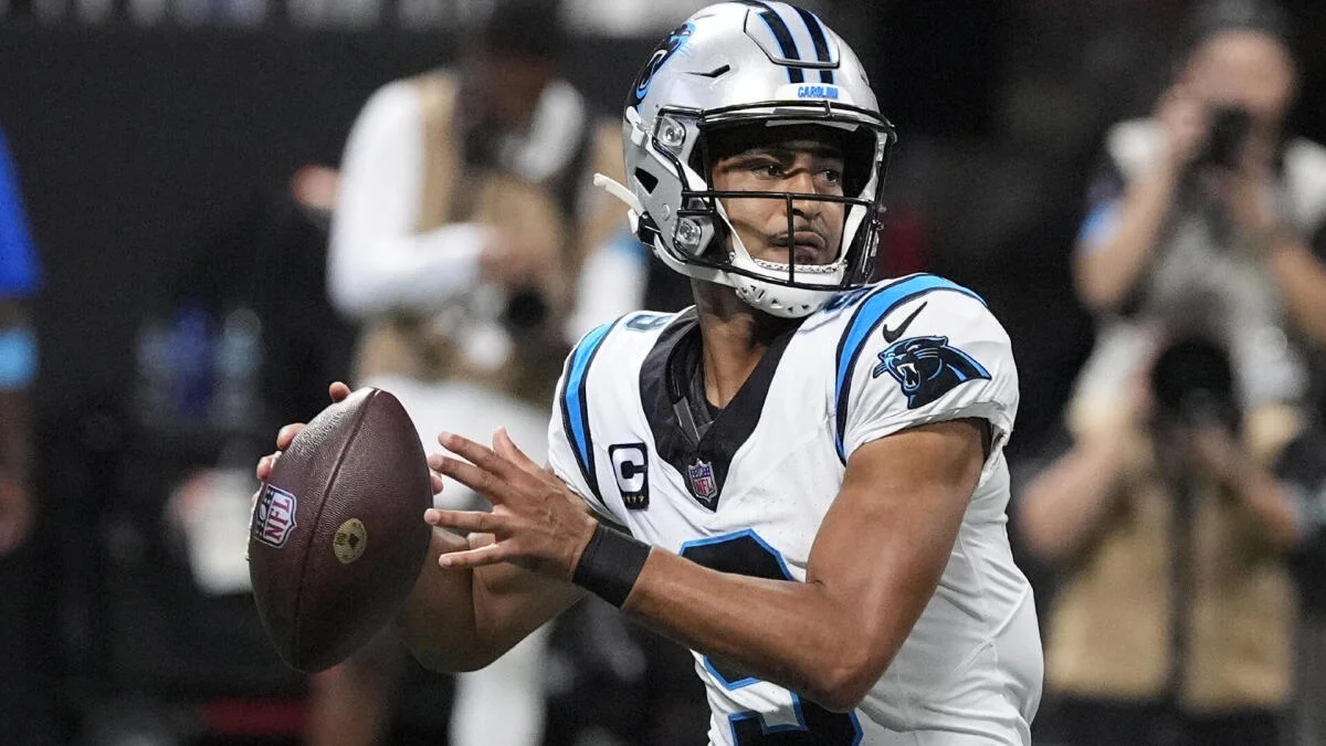 A Carolina Panthers quarterback in a white uniform holds a football, preparing to pass during a game.