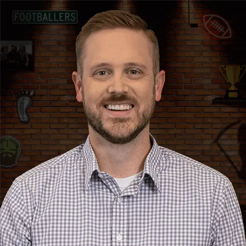Smiling bearded man in a checkered shirt poses in front of a football-themed brick wall.