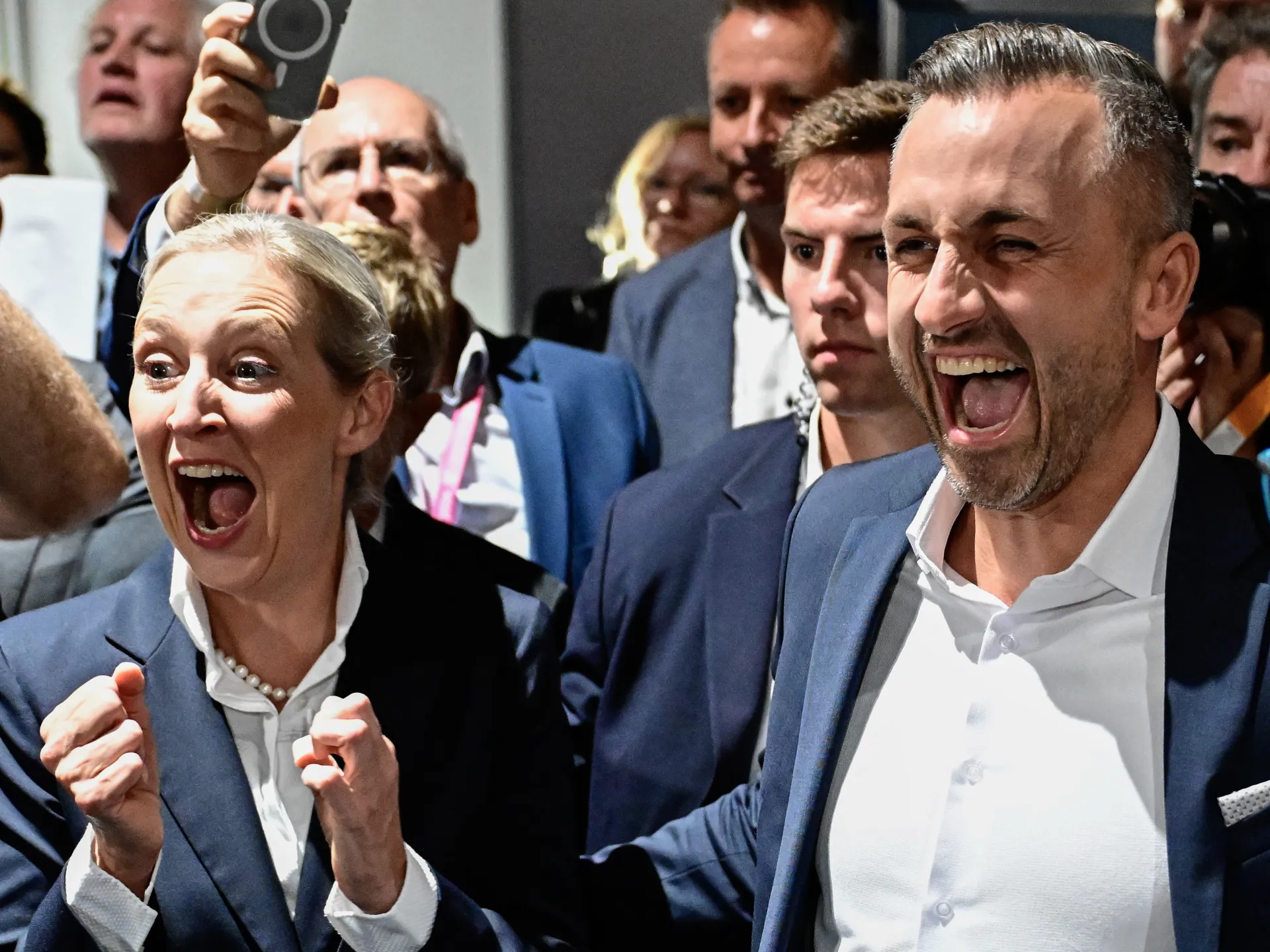 Two jubilant people in suits react excitedly amid a crowded indoor gathering.