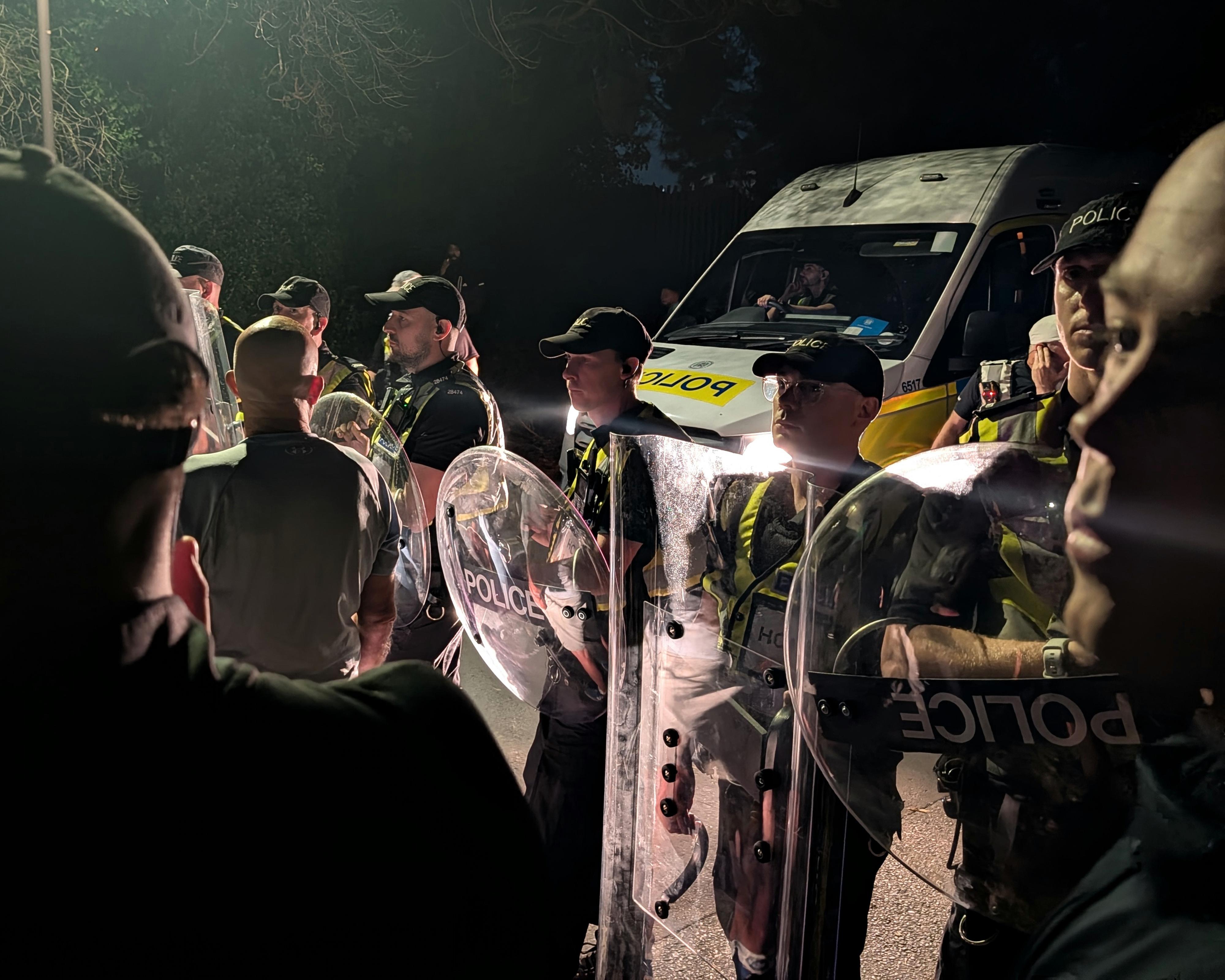 Police officers with riot shields stand beside a police van at night amid a crowd.