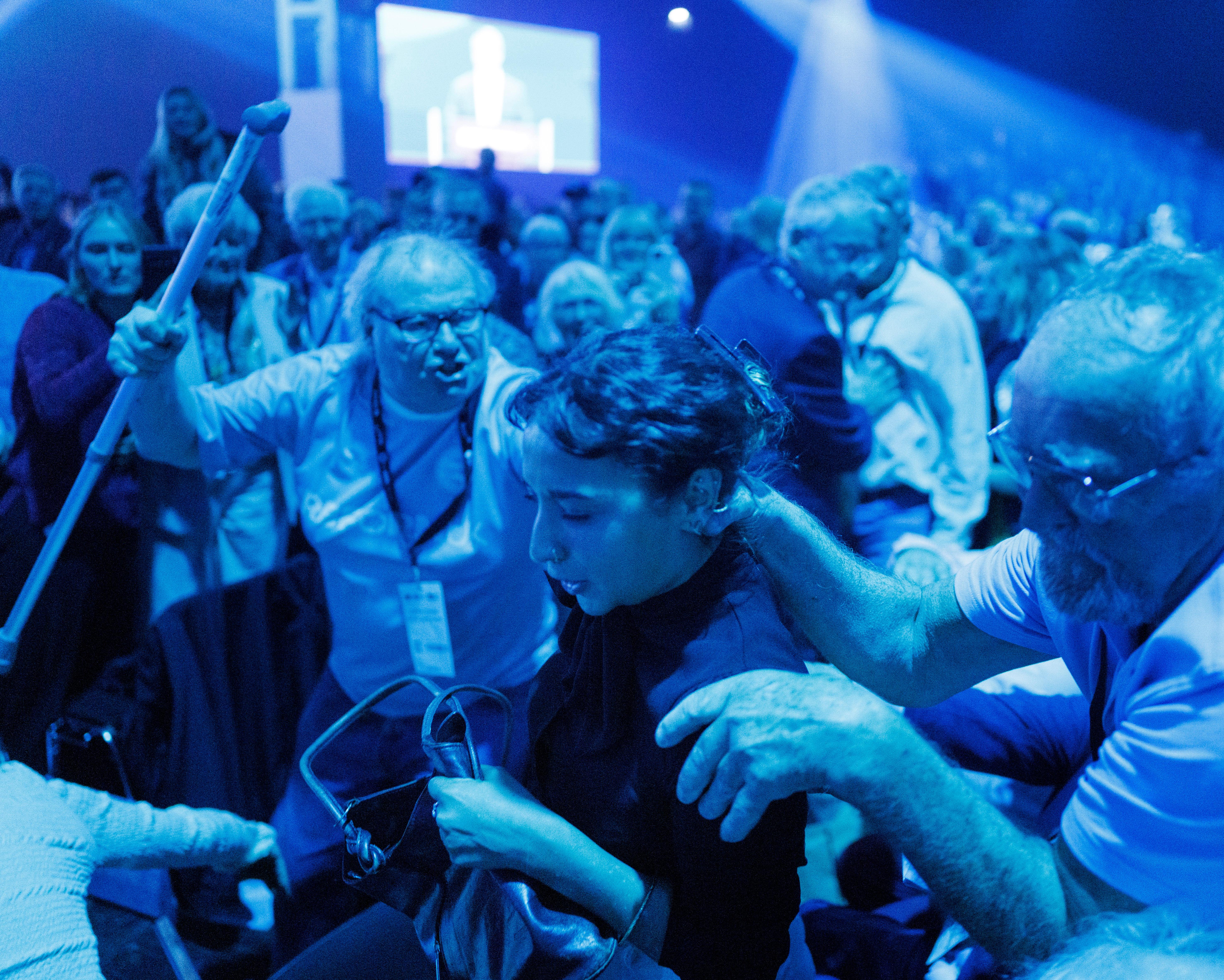 Crowd gathers under blue lighting as a person in front is assisted through the busy venue.