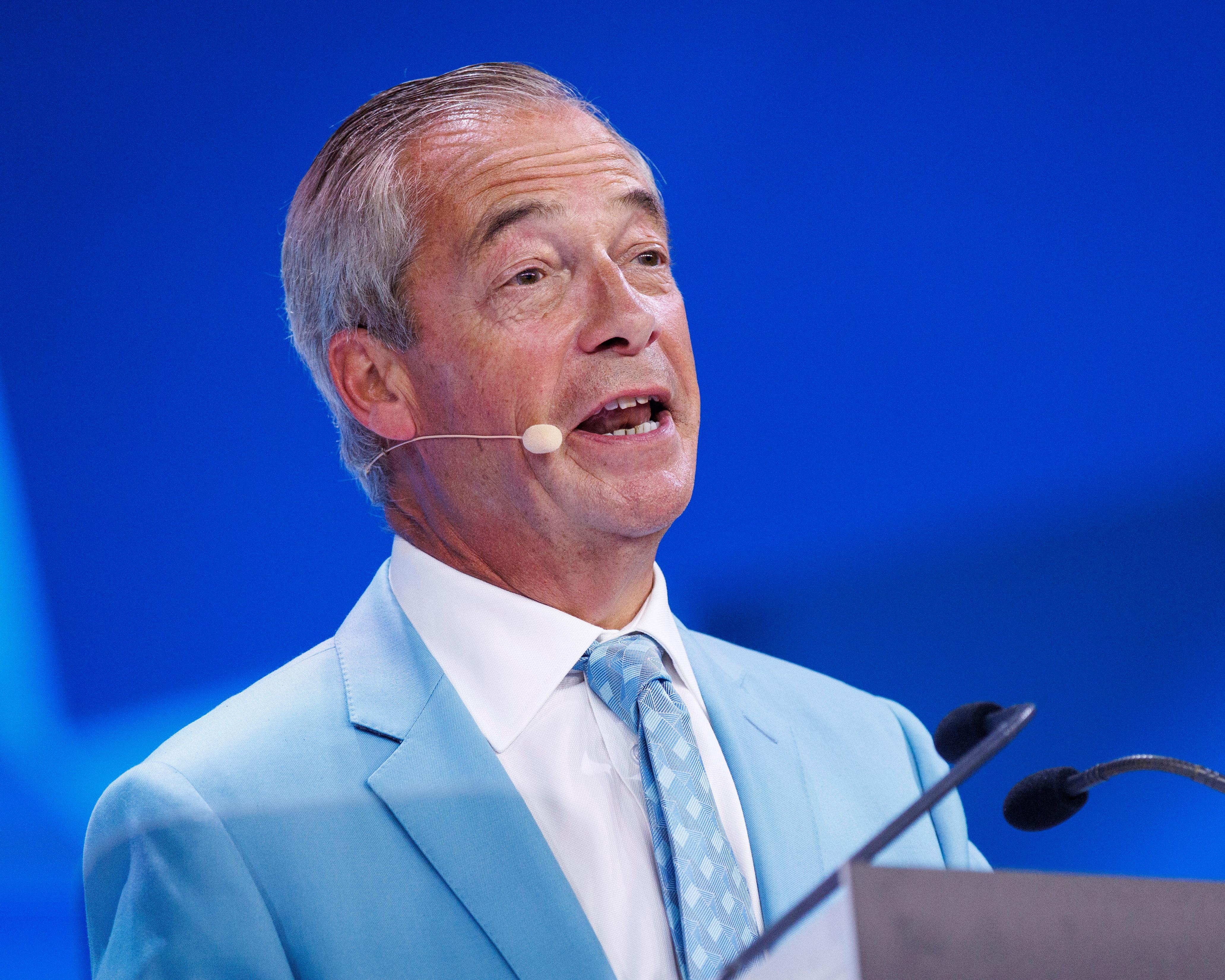 A grey-haired man in a pale blue suit speaks into a headset microphone.