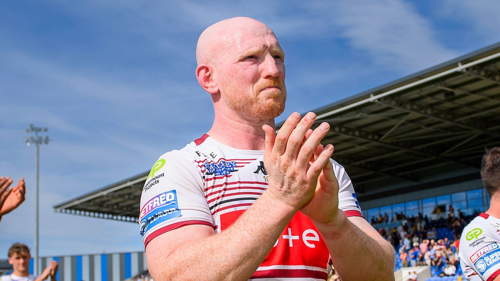 Rugby player in a red-and-white jersey applauds outdoors beneath a blue sky