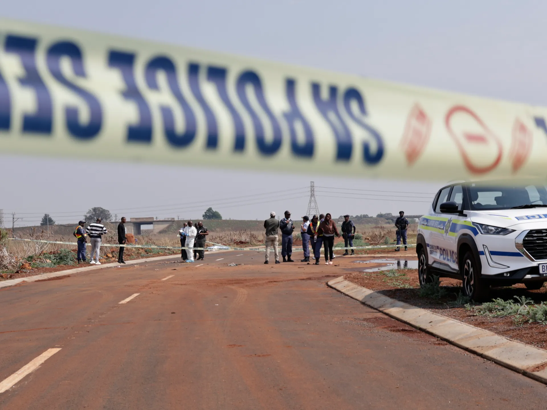 Police officers and bystanders gather behind crime-scene tape beside a police vehicle on a roadway.