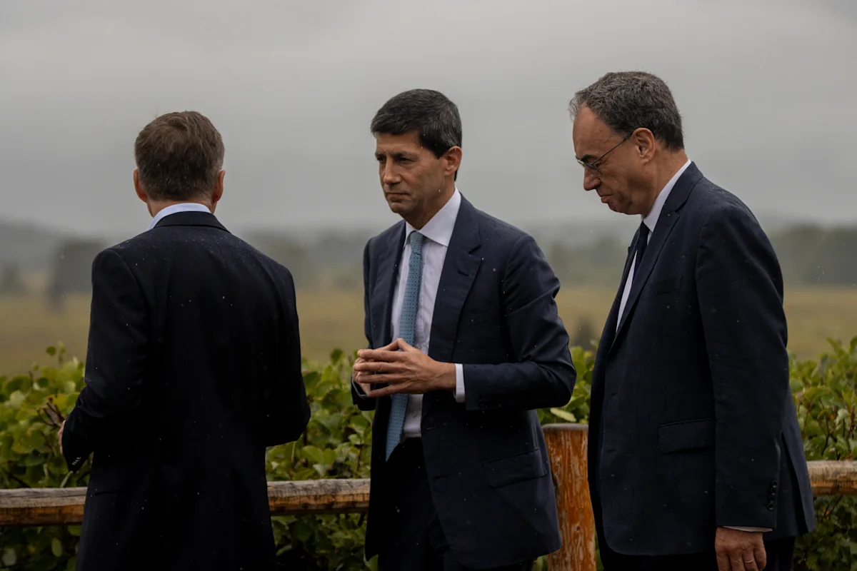 Three men in dark suits stand together outdoors near a vineyard during light rain.