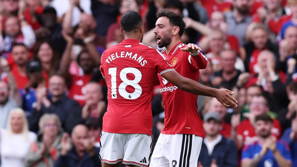 Two Manchester United players in red shirts celebrate in front of cheering supporters.