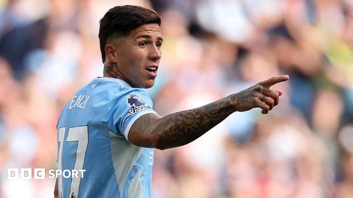 Footballer in a light blue jersey points during a match, with a blurred crowd behind him.