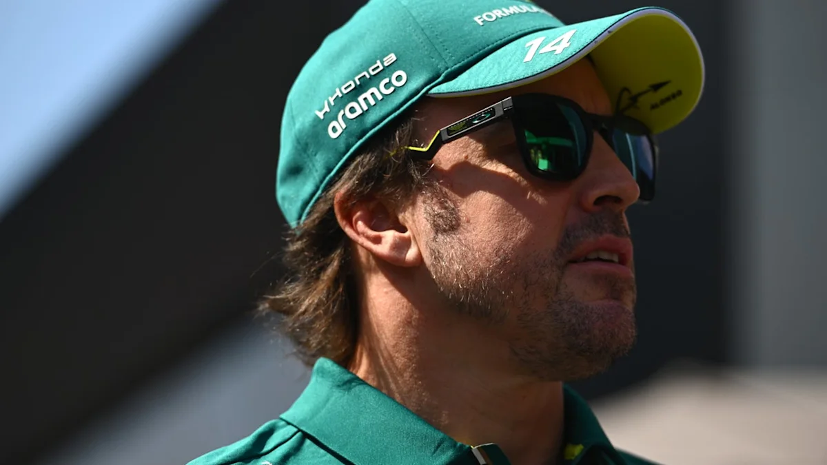 A man in a teal Aston Martin cap and shirt wears sunglasses in bright sunlight.