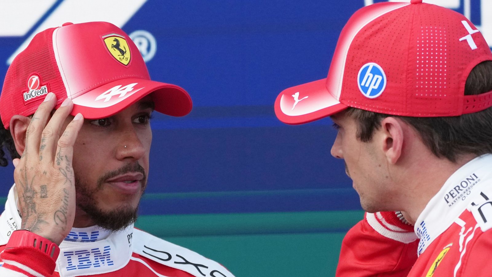 Two Ferrari drivers in red caps and uniforms speaking closely during a motorsport event