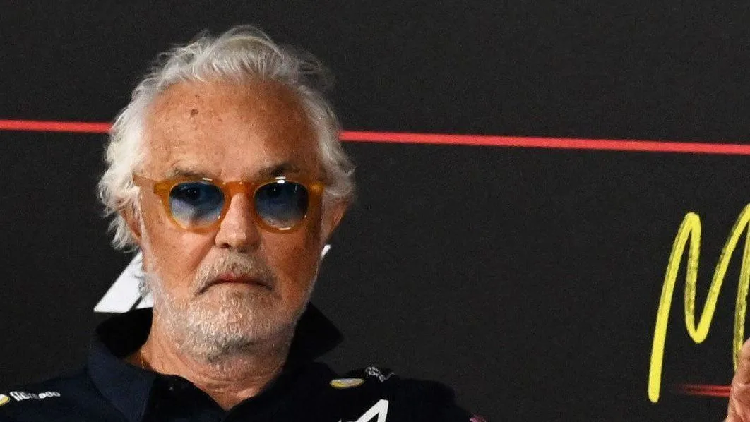 White-haired man wearing amber sunglasses and a black shirt against a dark branded backdrop