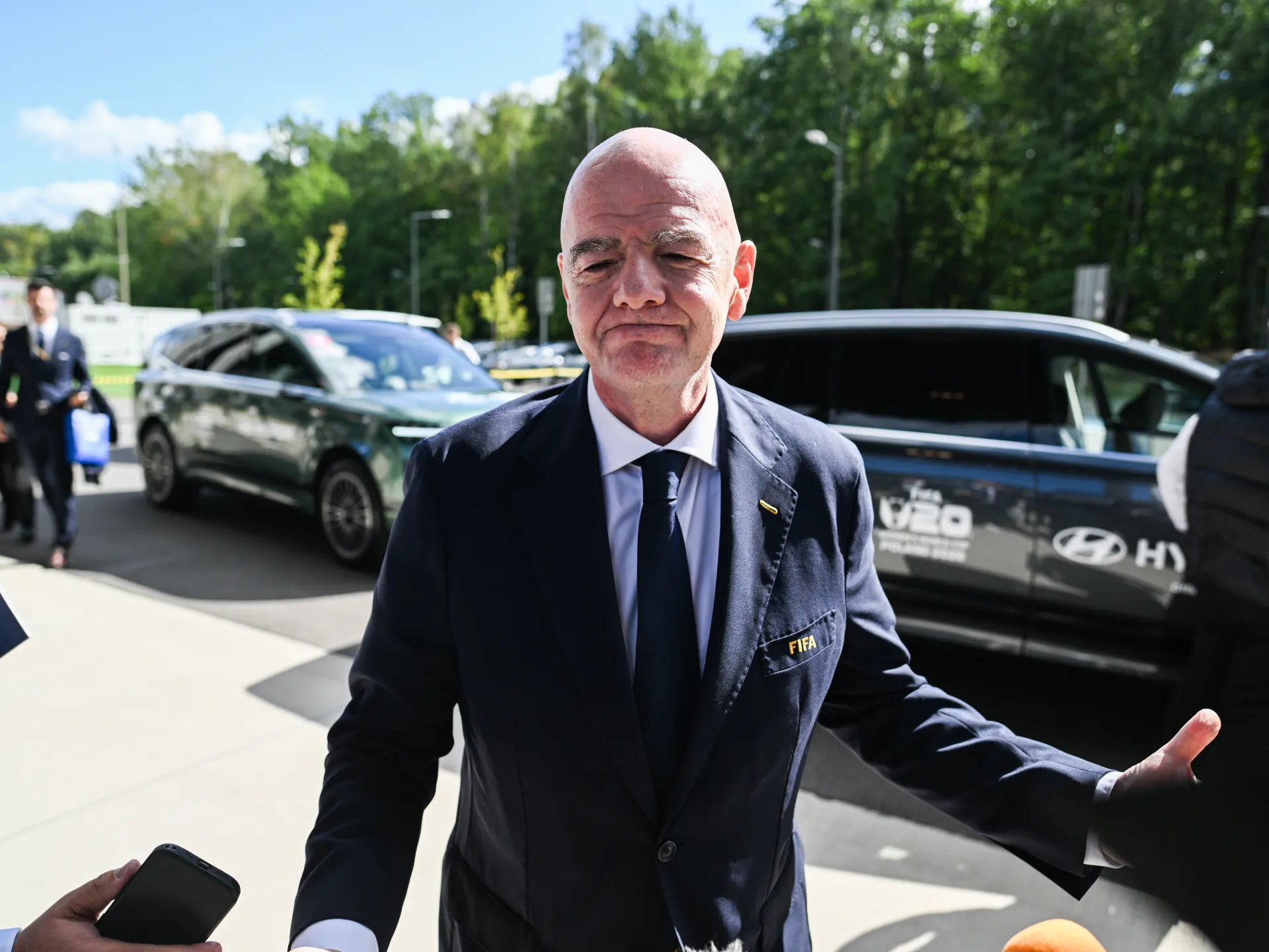 Bald man in a dark suit stands beside vehicles outdoors, extending one arm.