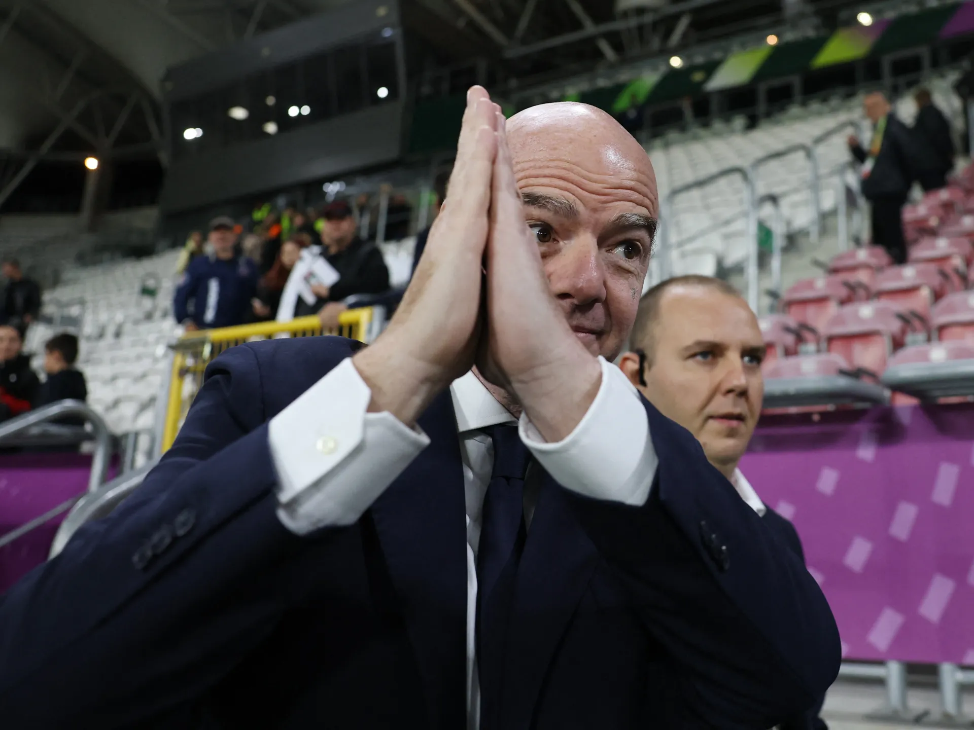 Bald man in a dark suit presses his palms together in a stadium stand.