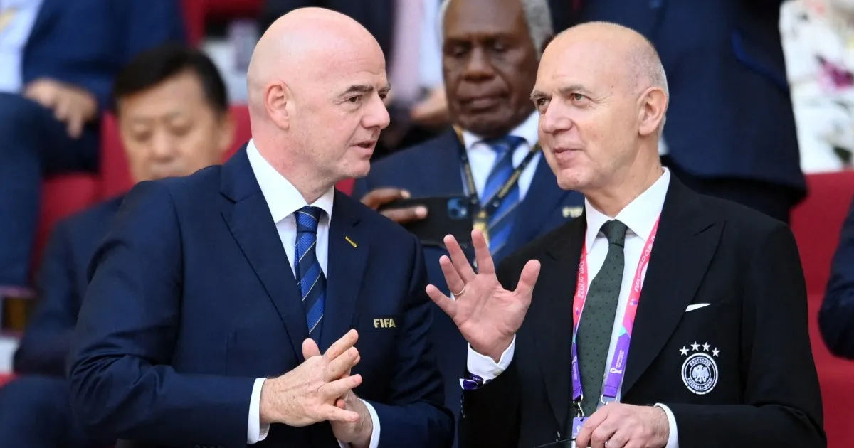 Two suited football officials converse courtside, one gesturing with an open hand.