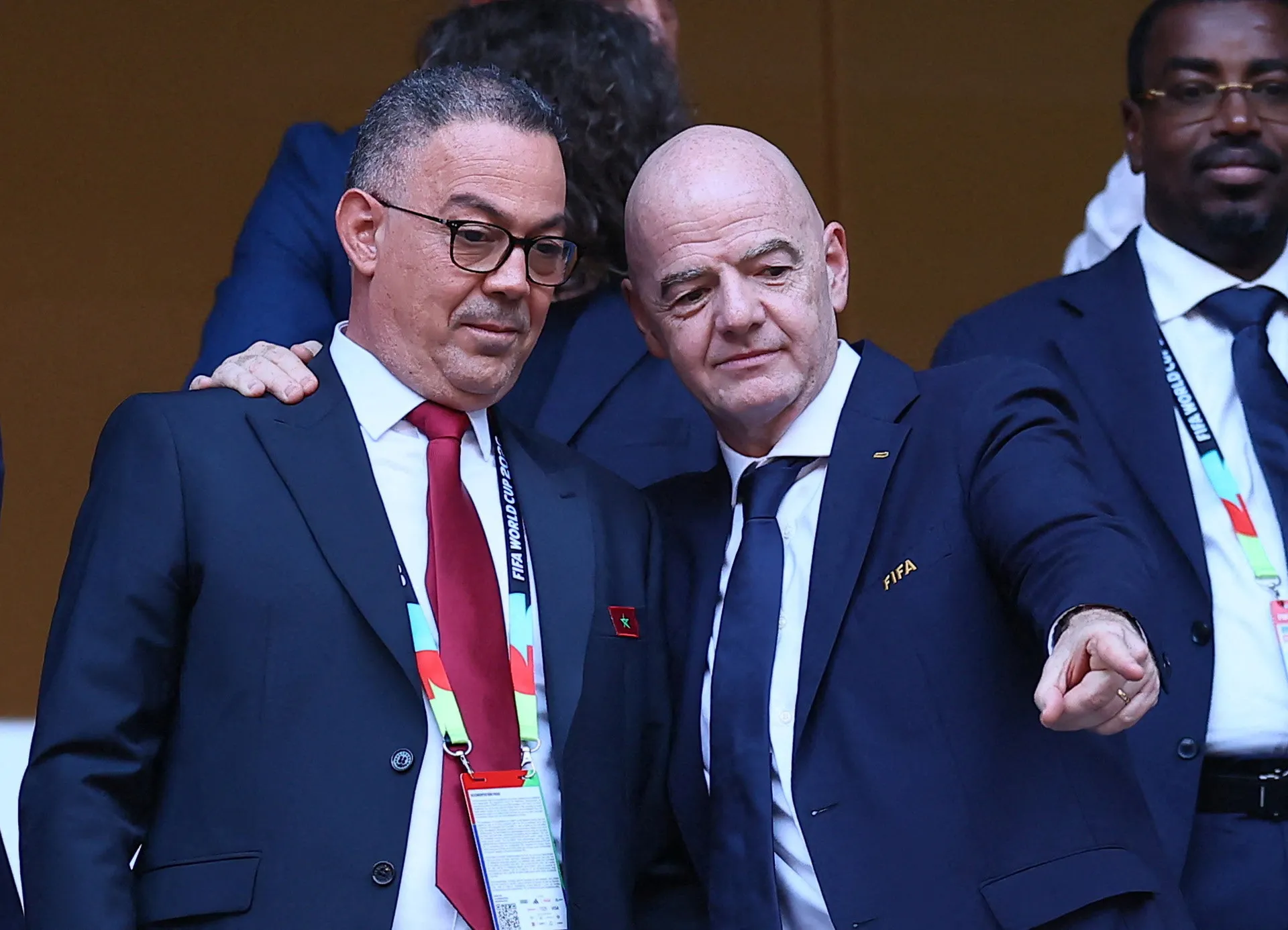 Two suited officials sit closely together at a FIFA event, with another delegate visible beside them.