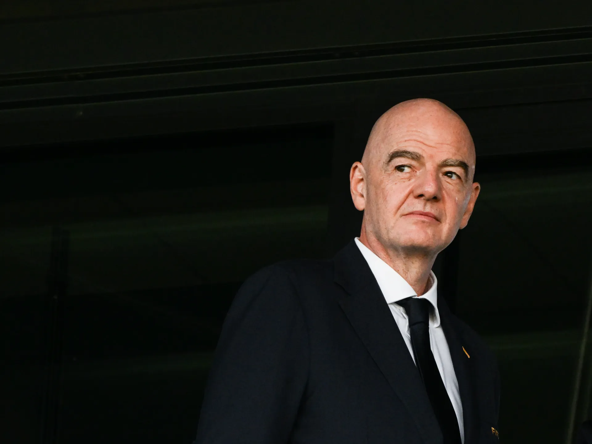 Bald man in dark suit and tie looks to the side against a dark background