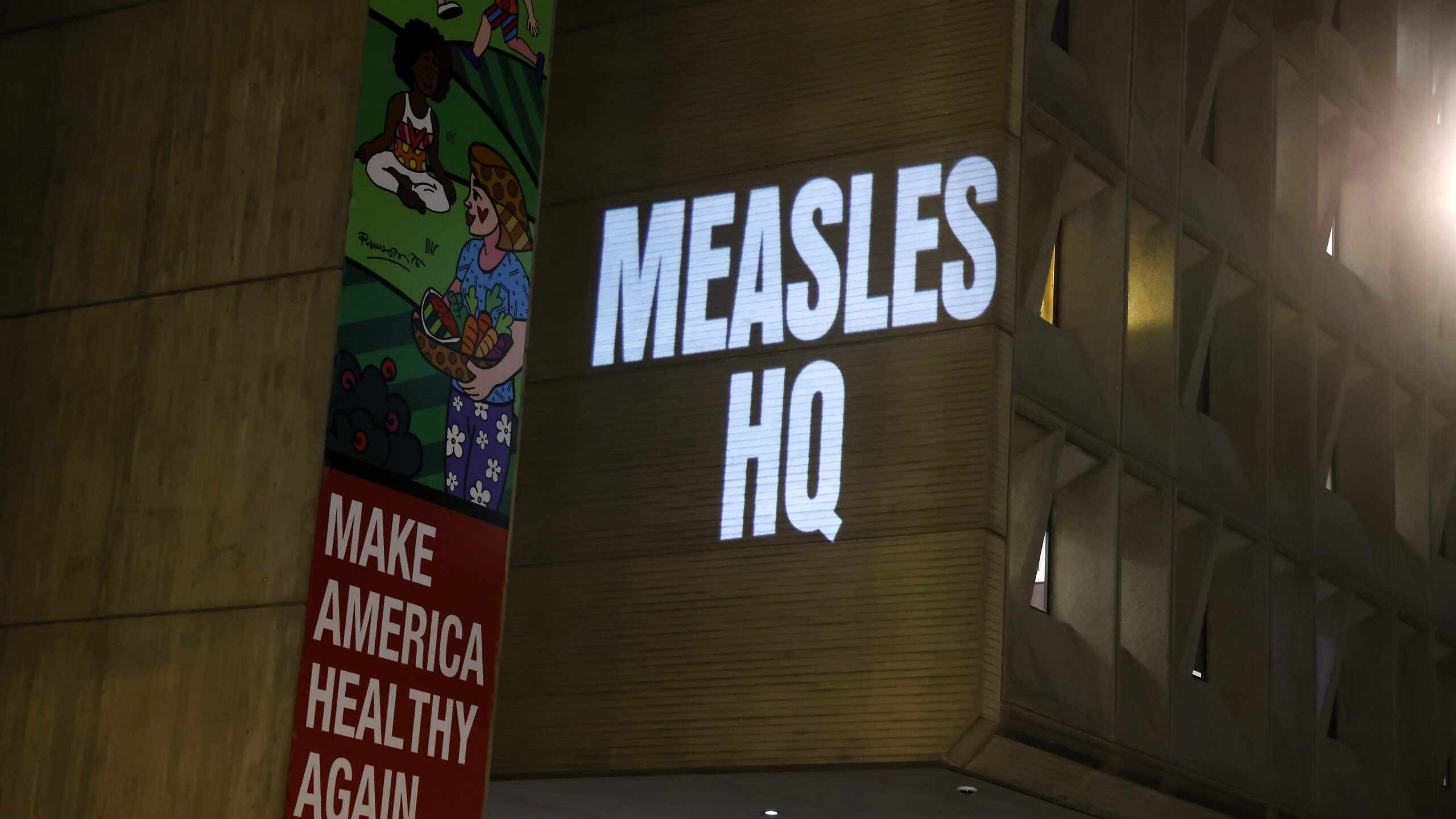 A projected white “MEASLES HQ” sign appears on a building beside a colorful health-themed banner.