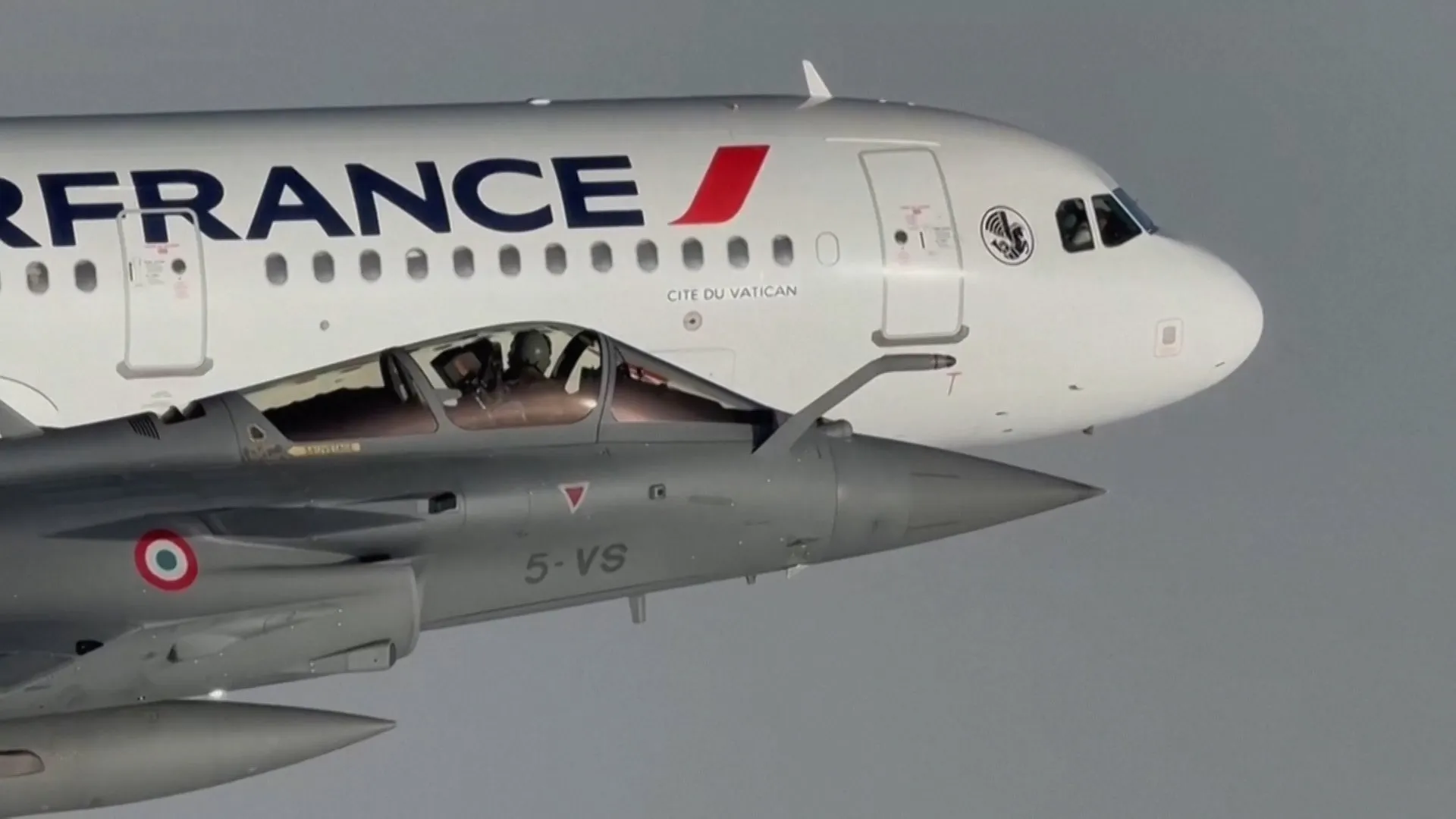 A gray fighter jet flies alongside an Air France passenger plane bearing Vatican City markings.