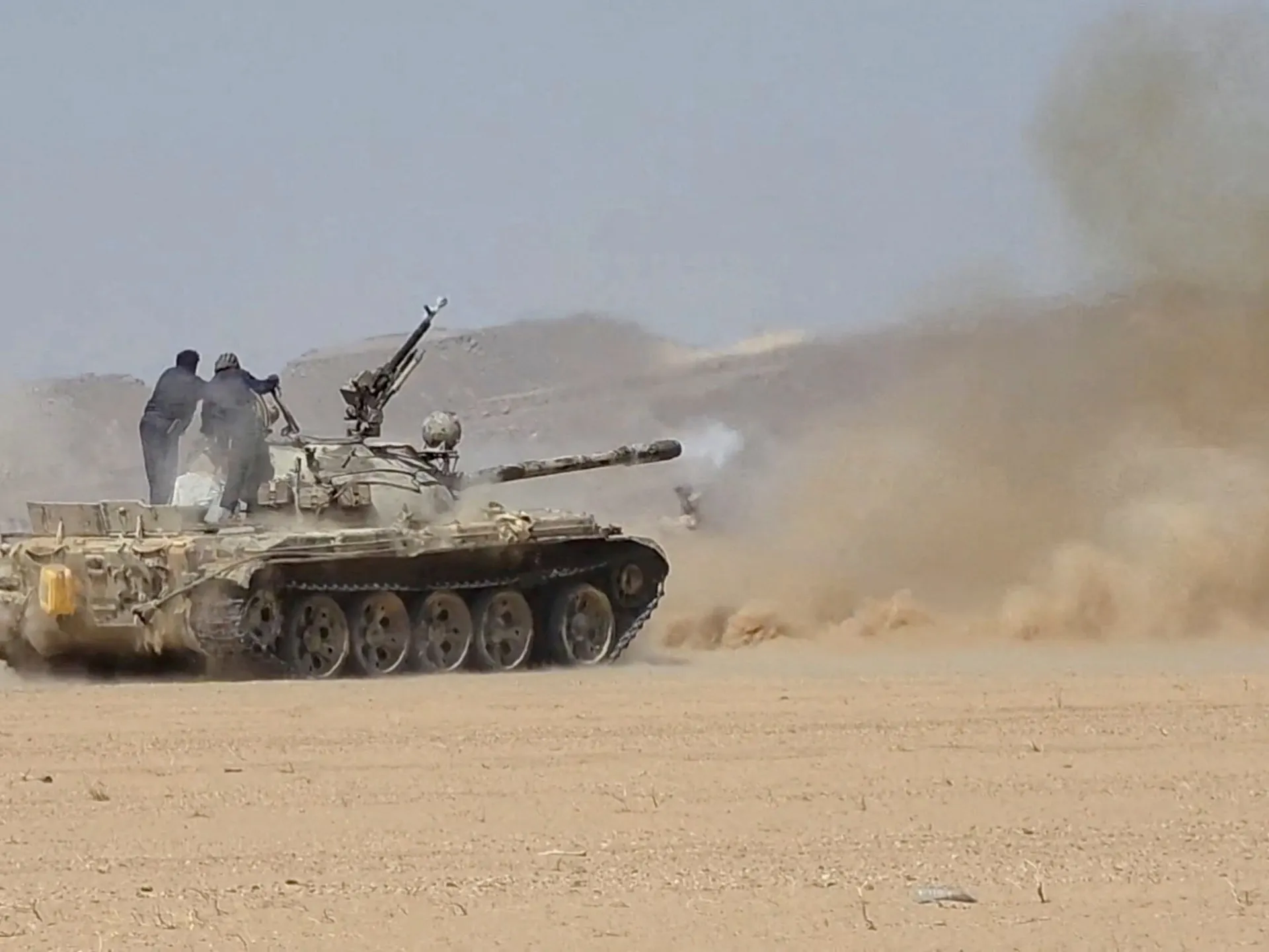 Armored tank firing across a dusty desert while two people stand on its turret