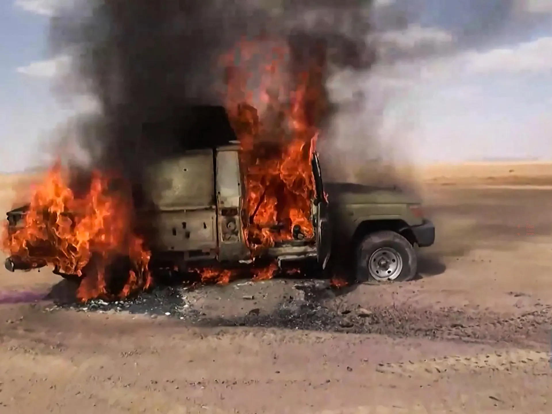 A pickup truck burns intensely on a desert roadside, sending thick black smoke into the sky.