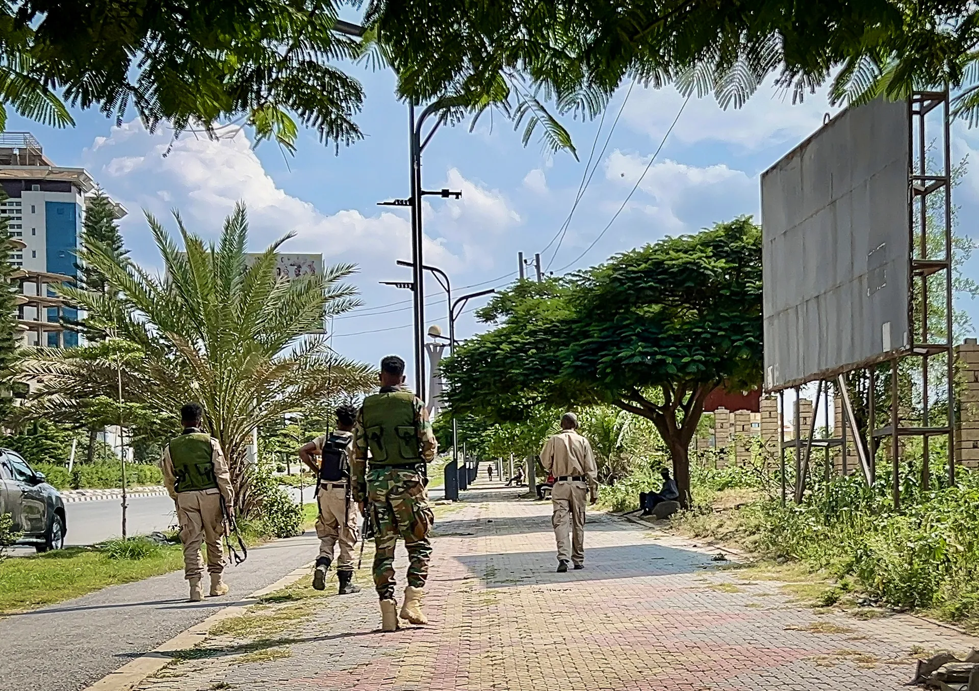 Three uniformed men walk along a tree-lined urban sidewalk beside buildings and a parked vehicle.