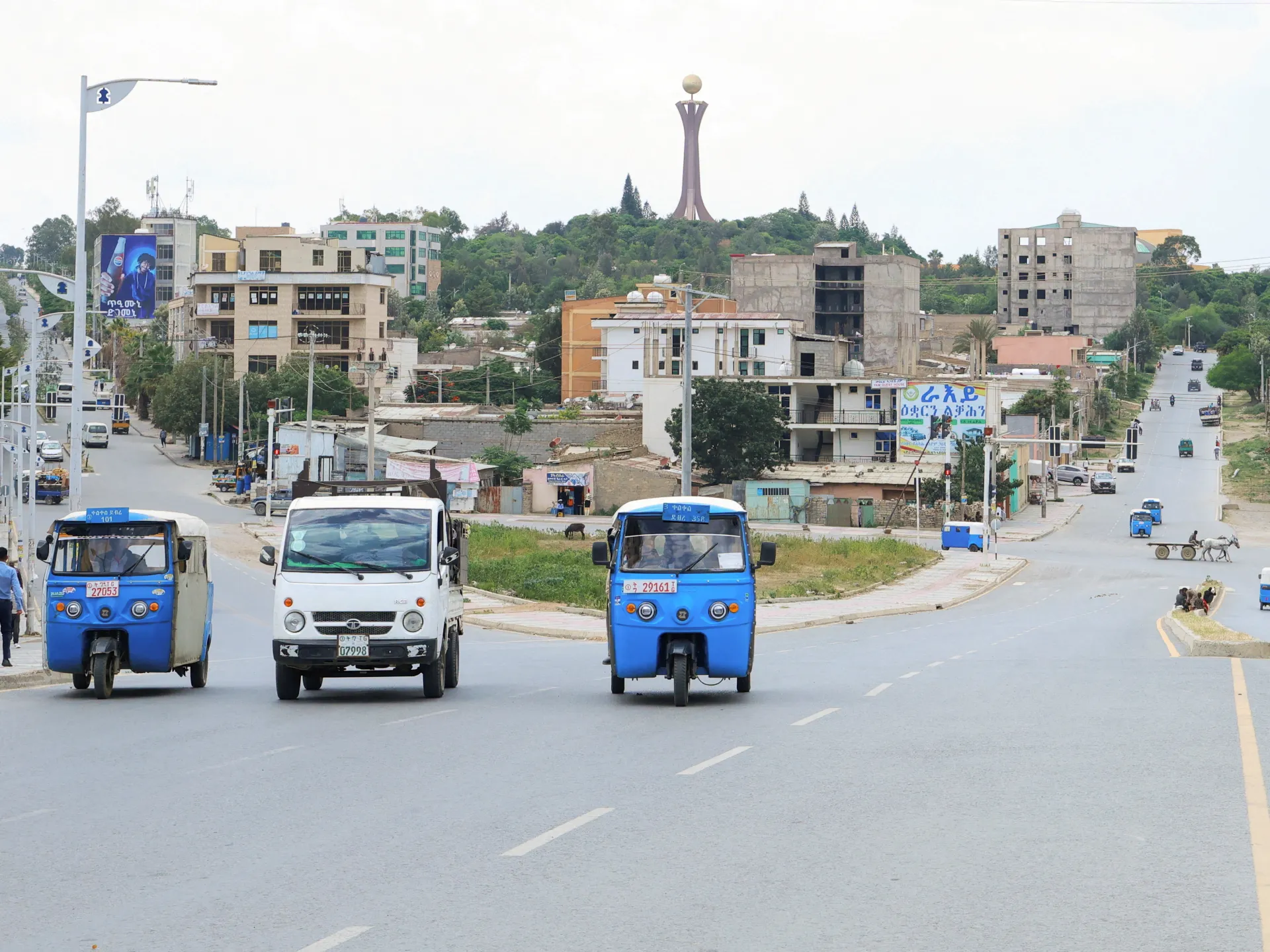 Three small vehicles travel along a broad urban road lined with buildings and trees.