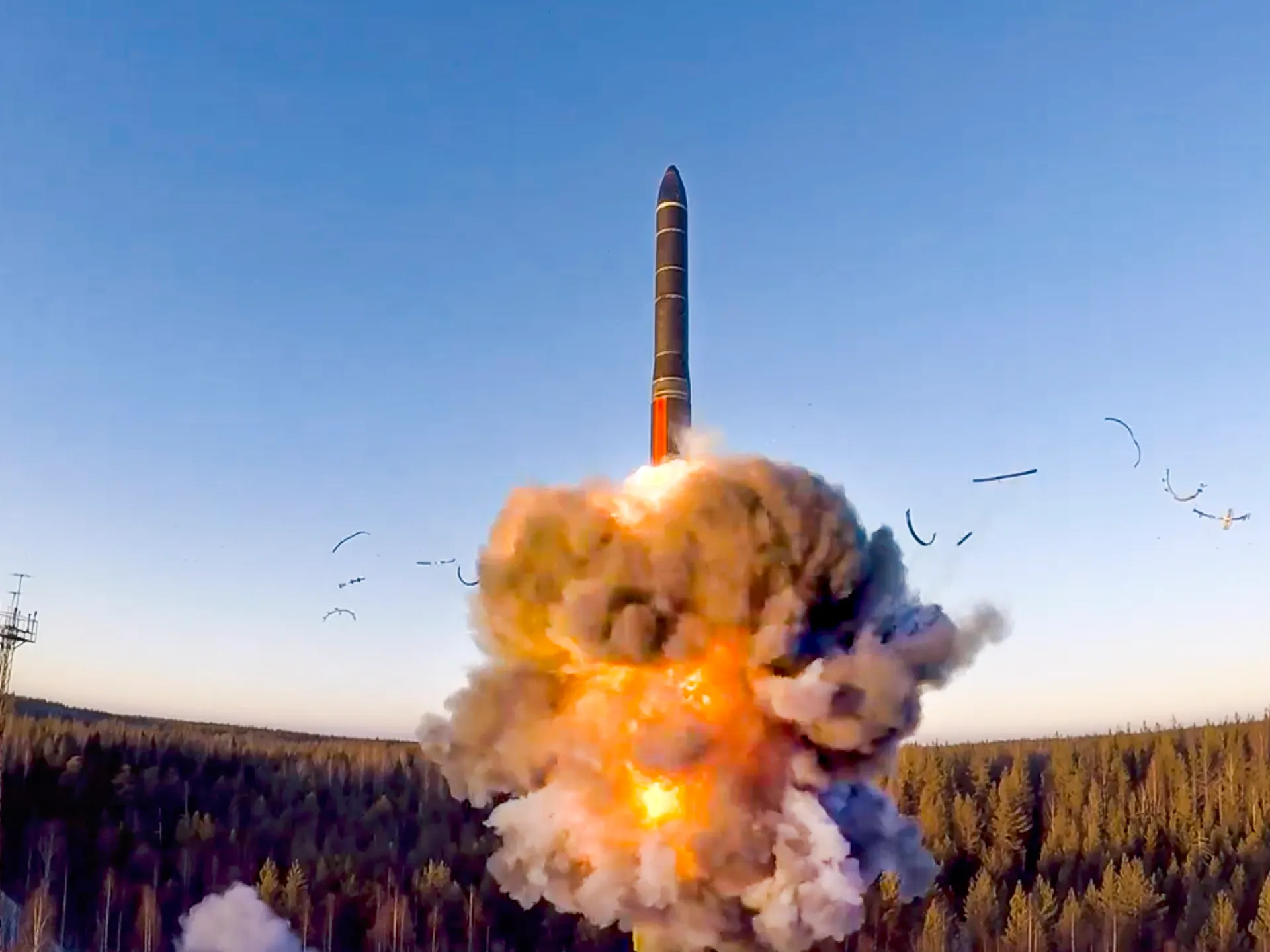 Rocket launches above a forest as a large orange fireball and smoke cloud billow outward.