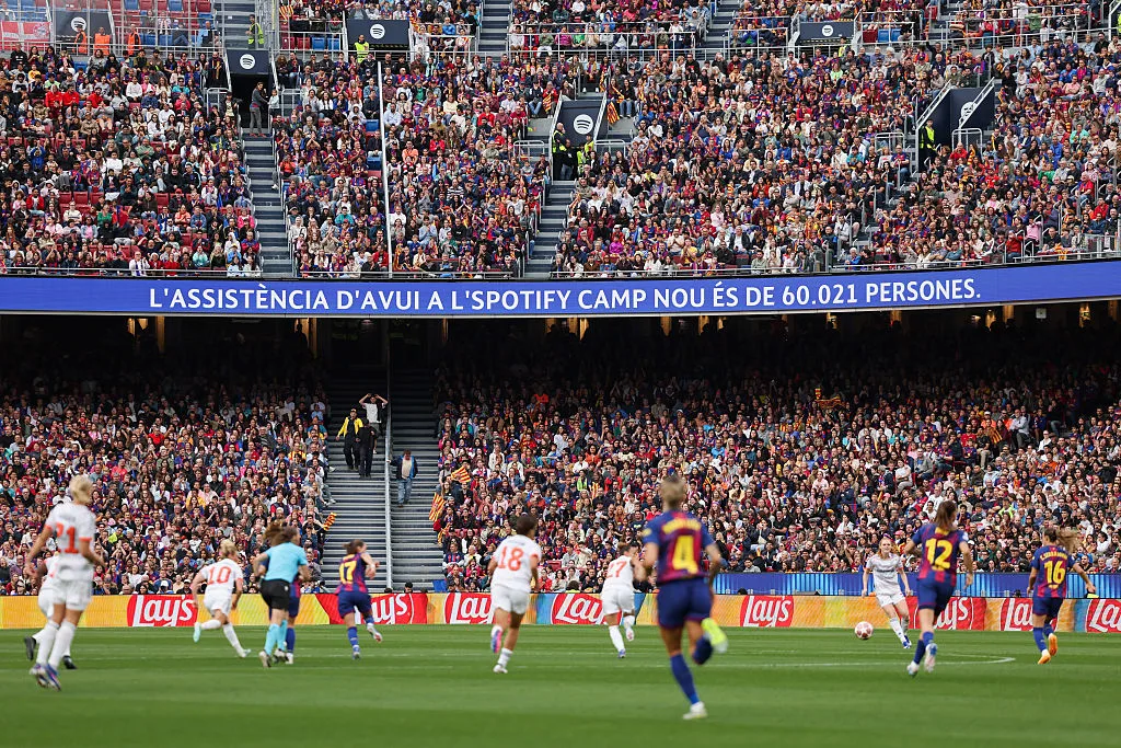 Footballers play before a packed stadium crowd, with a Catalan attendance message displayed above the stands.