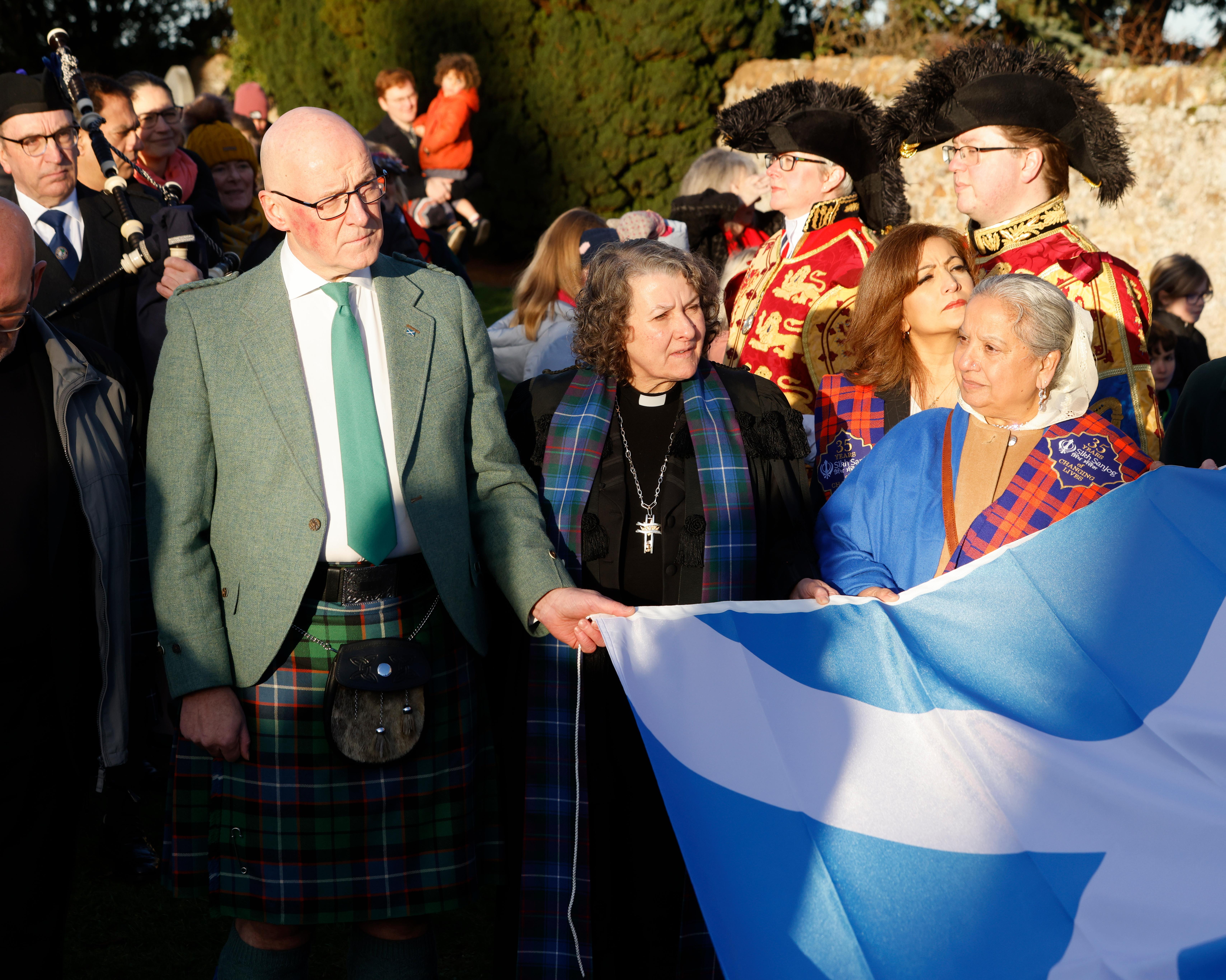 People in formal and traditional Scottish attire hold a blue-and-white flag outdoors.