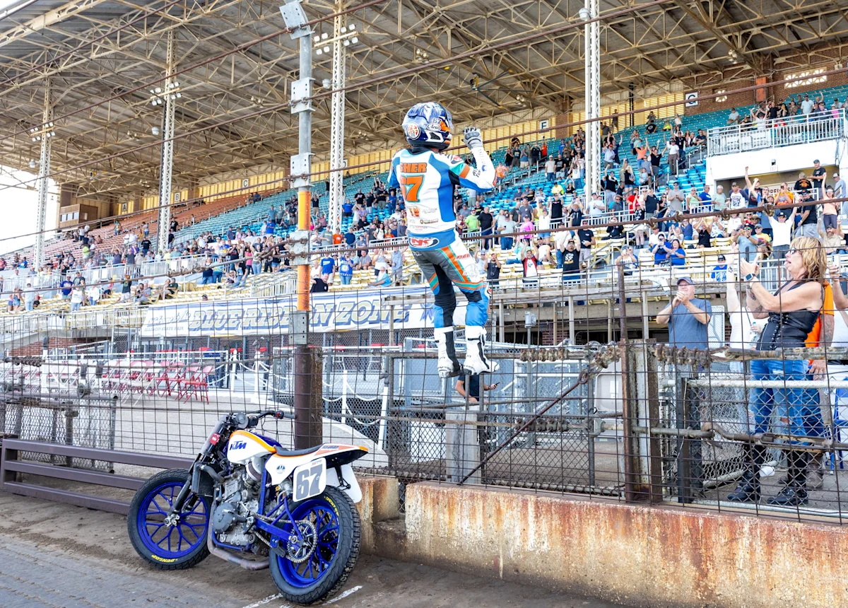 Motorcycle racer raises a hand beside a numbered bike as spectators watch from the stands.